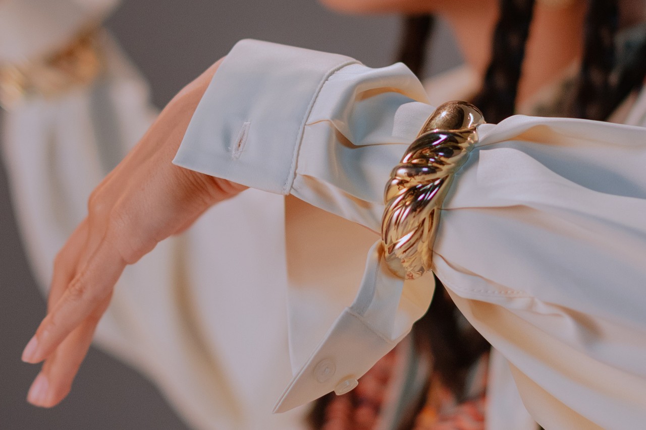 a woman’s extended arm wearing a white shirt sleeve and a chunky gold bracelet