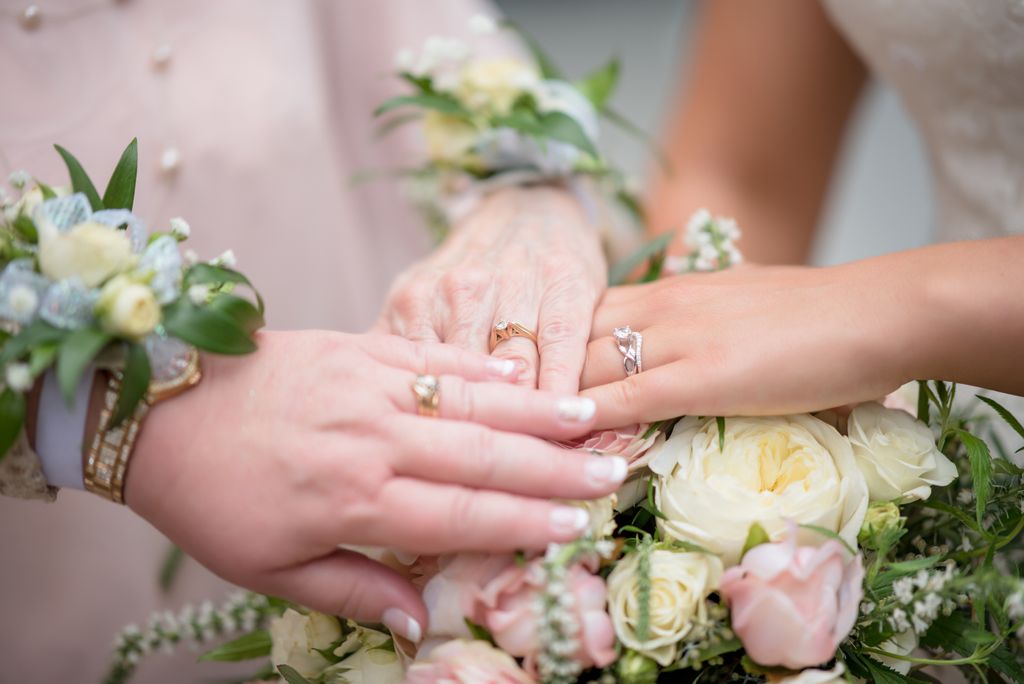 Symbolizing generational connection, a close-up of three women’ hands resting on a bouquet of pink and white roses, with emphasis on their wedding rings.