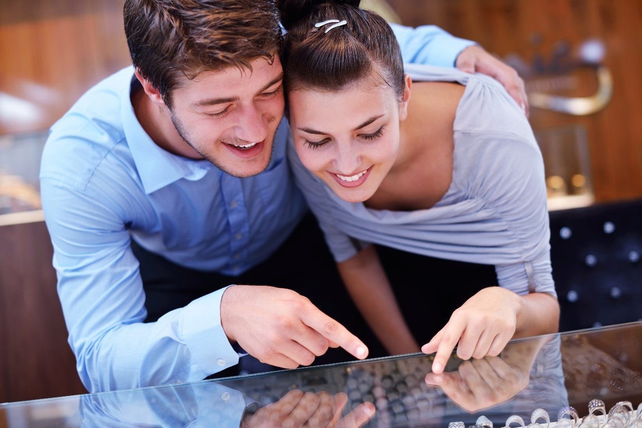 A smiling couple leans over a jewelry store display, pointing at engagement rings.