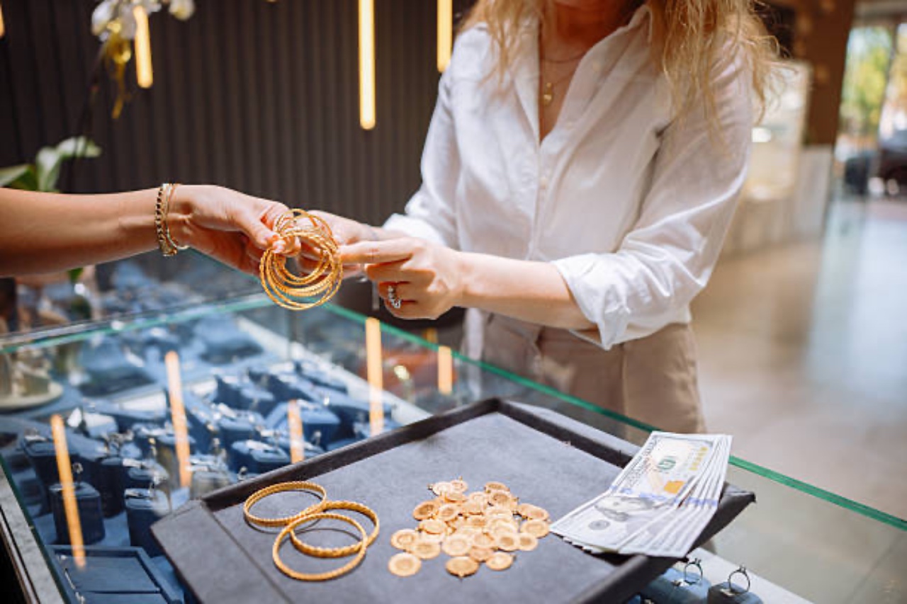 A jeweler showing gold bangles to a customer over a display case with cash and gold coins on a tray.