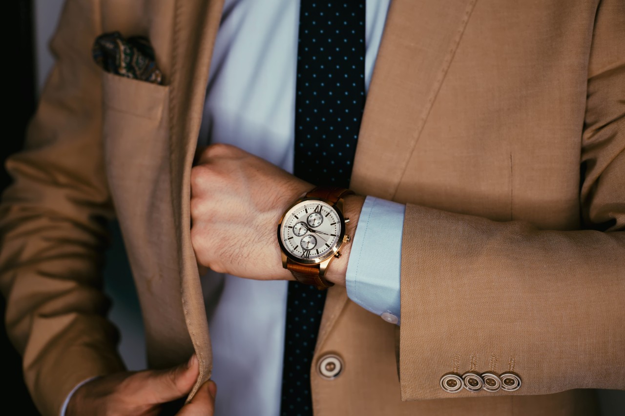 A close up of a man in a beige suit, showcasing a brown leather wristwatch with a beige dial.