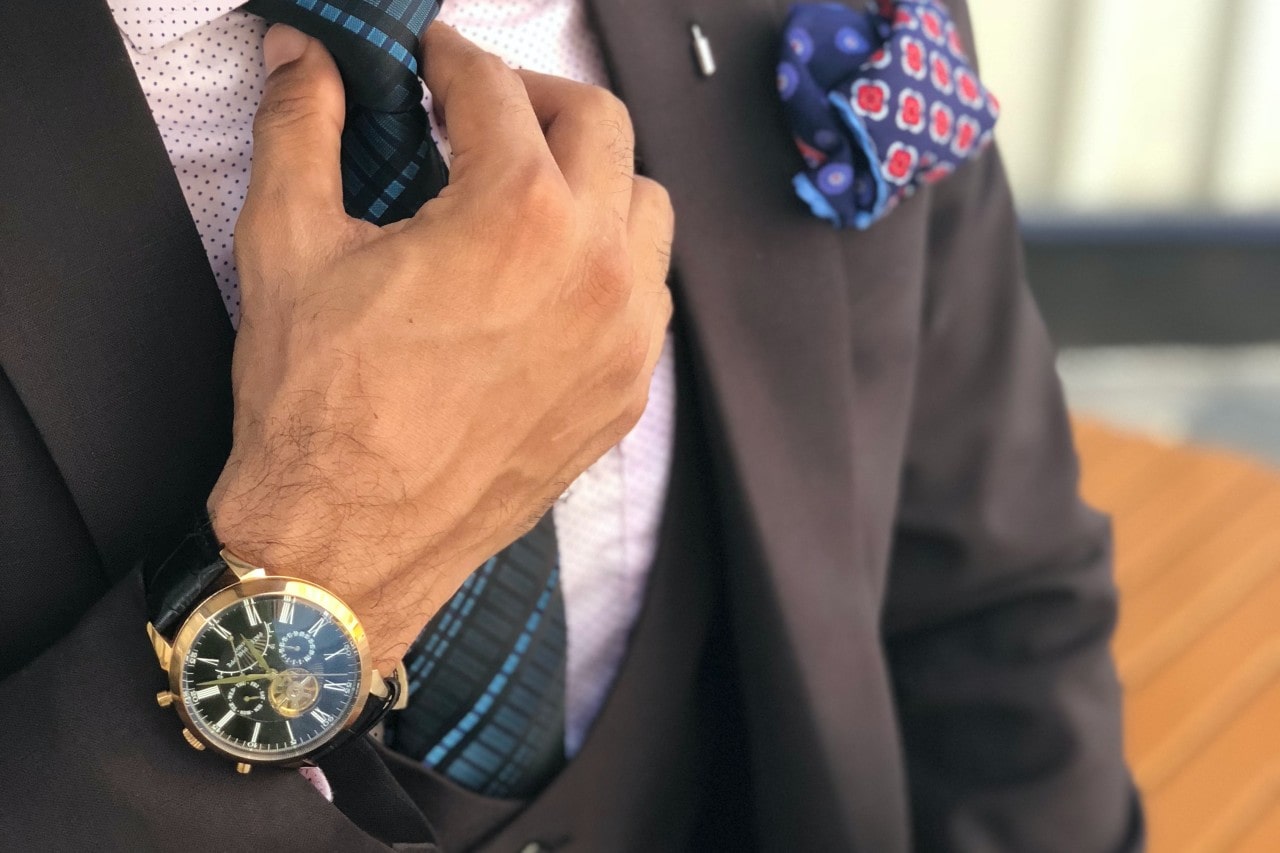 A close up of a guy in a brown suit fixing his tie, presenting a black large wristwatch.
