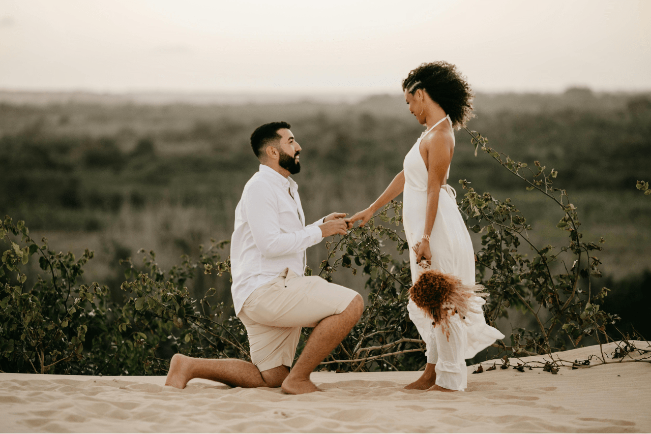 A man kneels to propose to a woman in a white dress holding dried flowers on a sandy area with bushes in the background.