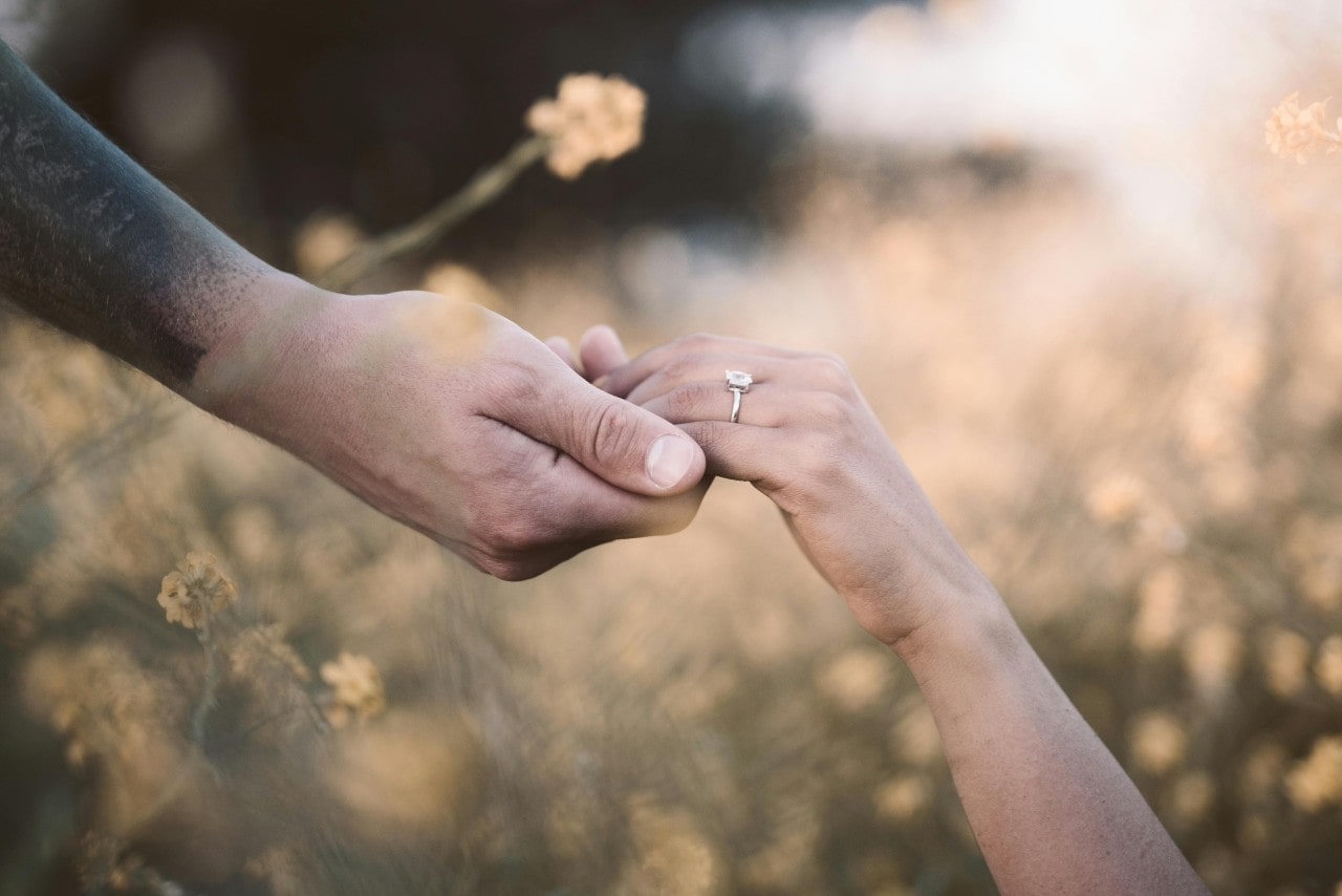 Close-up of a couple holding hands in a wildflower field, with a strong emphasis on a diamond engagement ring on one hand.
