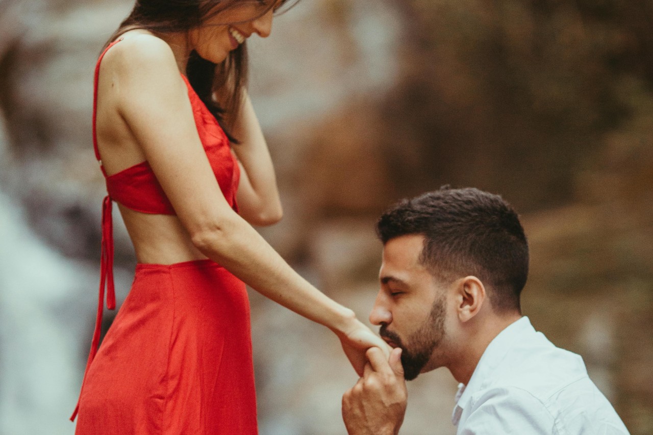A man in a white shirt kneels to kiss a woman's hand while proposing outdoors. She wears a red dress.