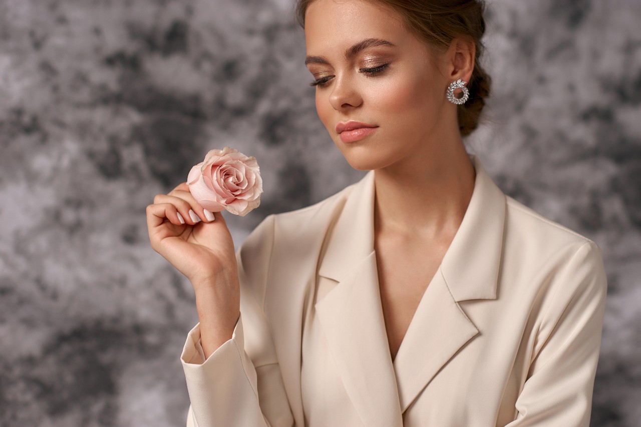 A woman in beige blaze holds a pink rose in her hand, showcasing round diamond stud earring.