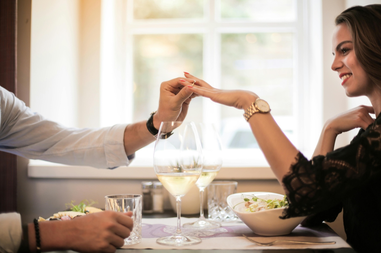A man putting an engagement ring on a woman’s hand, while she wears a yellow gold wristwatch and black lace blouse, all while sitting on a dining table at a restaurant.