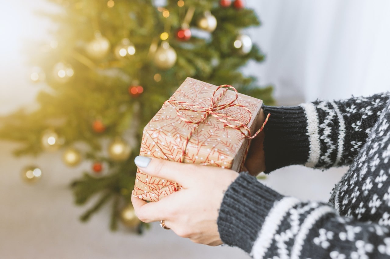 A close up of a person in a grey sweater holding a small giftbox against a blurry background of a christmas tree