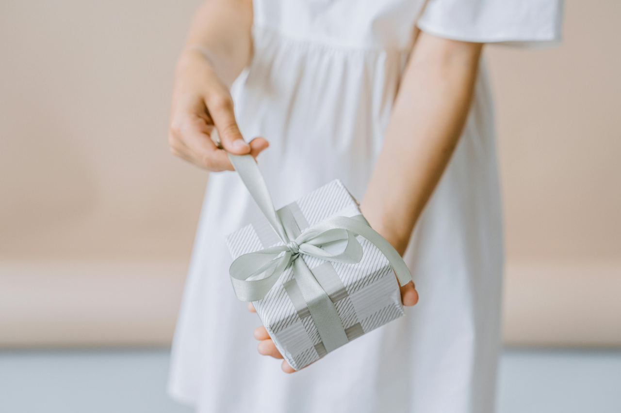 A child in a white dress holds out a small striped gift box tied with a satin ribbon.