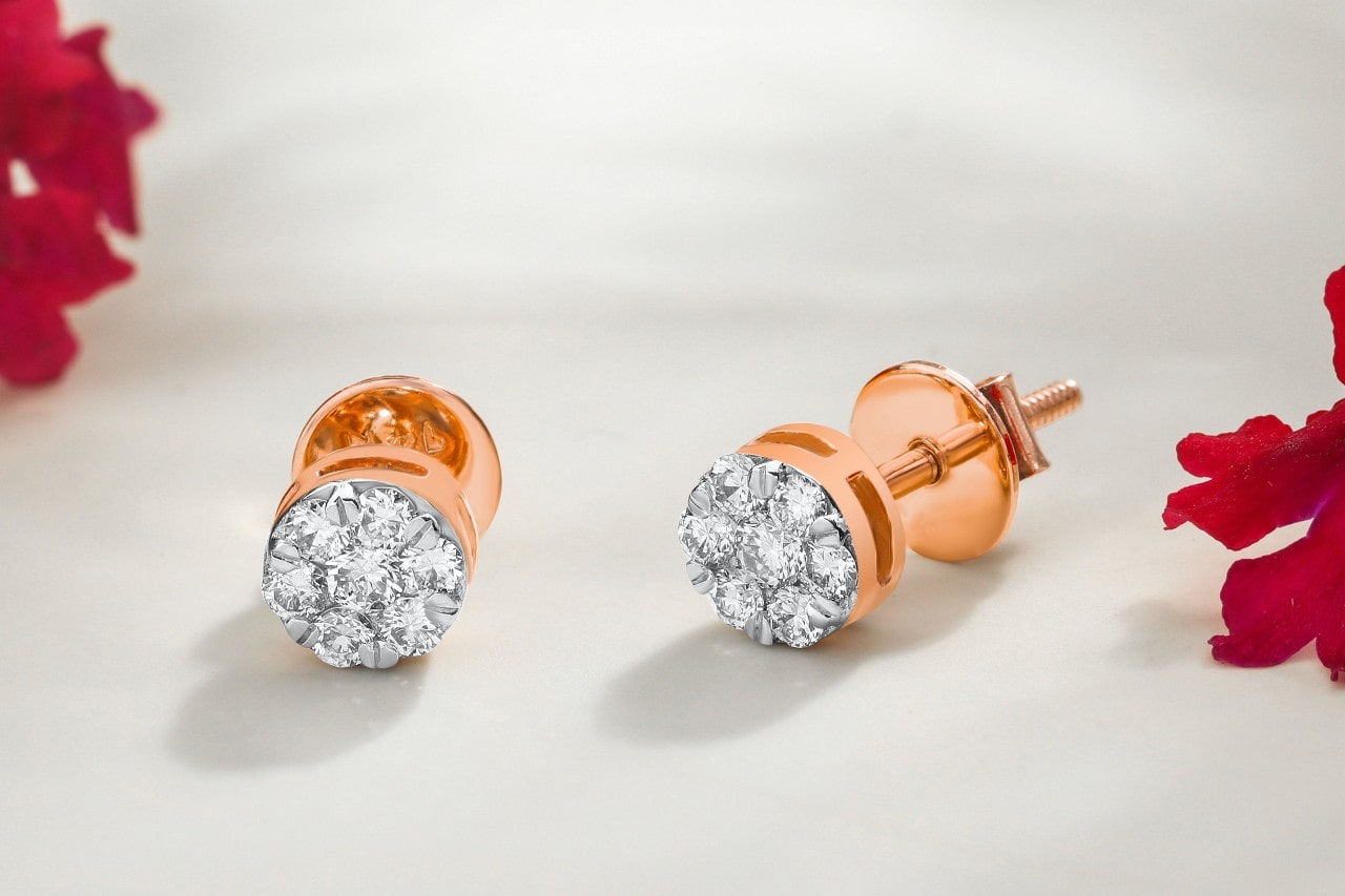 A pair of rose gold diamond stud earrings on a white surface, red flowers laying on the table around them.
