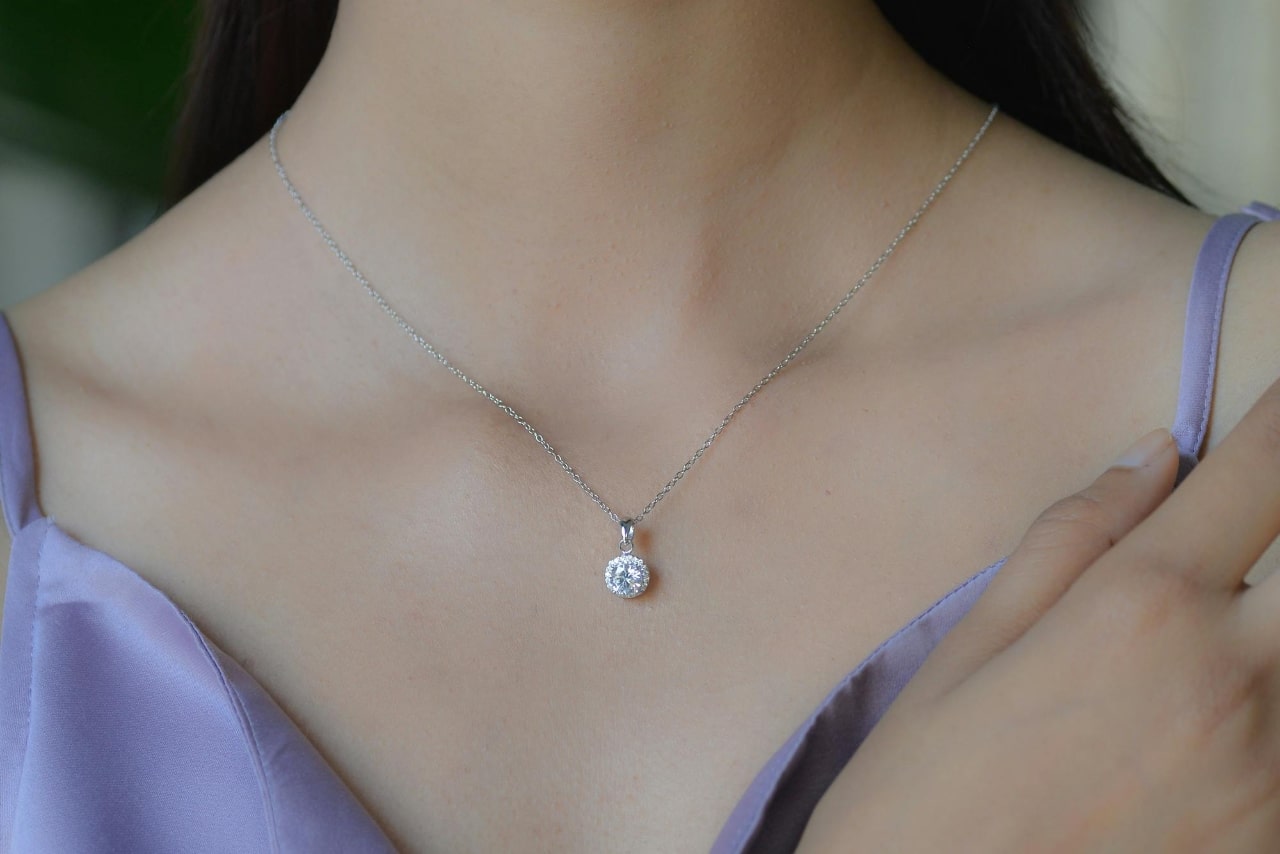 A close up of a white gold diamond pendant on a woman’s neck, dressed in a lavender satin top