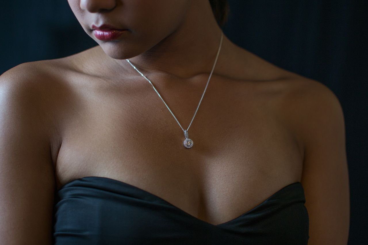 A woman wearing a strapless black dress and a delicate diamond pendant stands against a dark background.