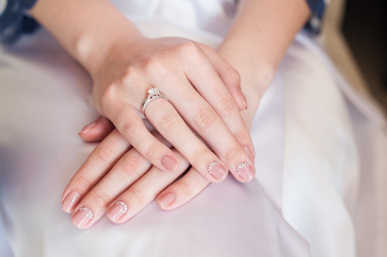 Close-up of hands wearing a silver engagement ring resting on soft white fabric, with light pink manicured nails accented by subtle rhinestones.