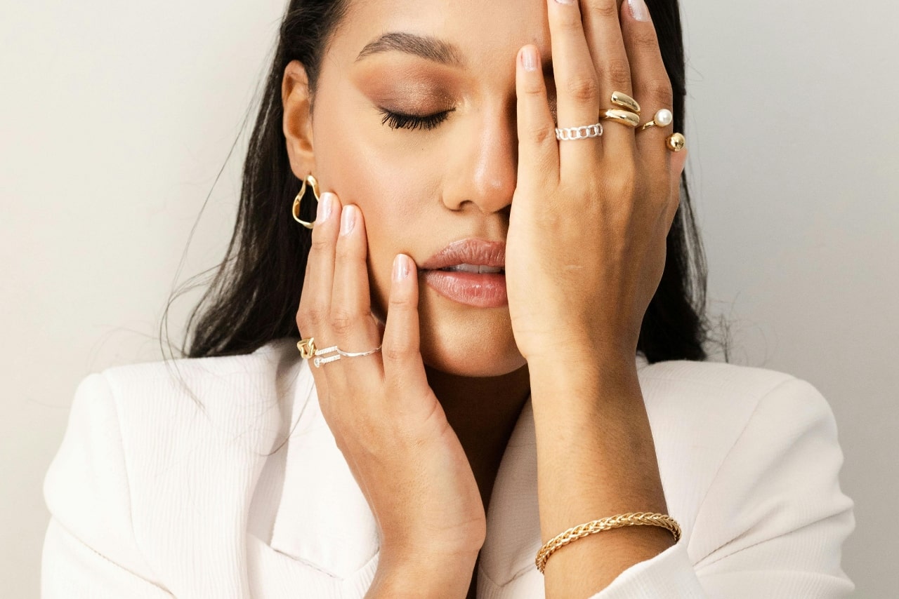 A woman in a white blazer, eyes closed, covers one eye with her hand adorned with stylish jewelry.