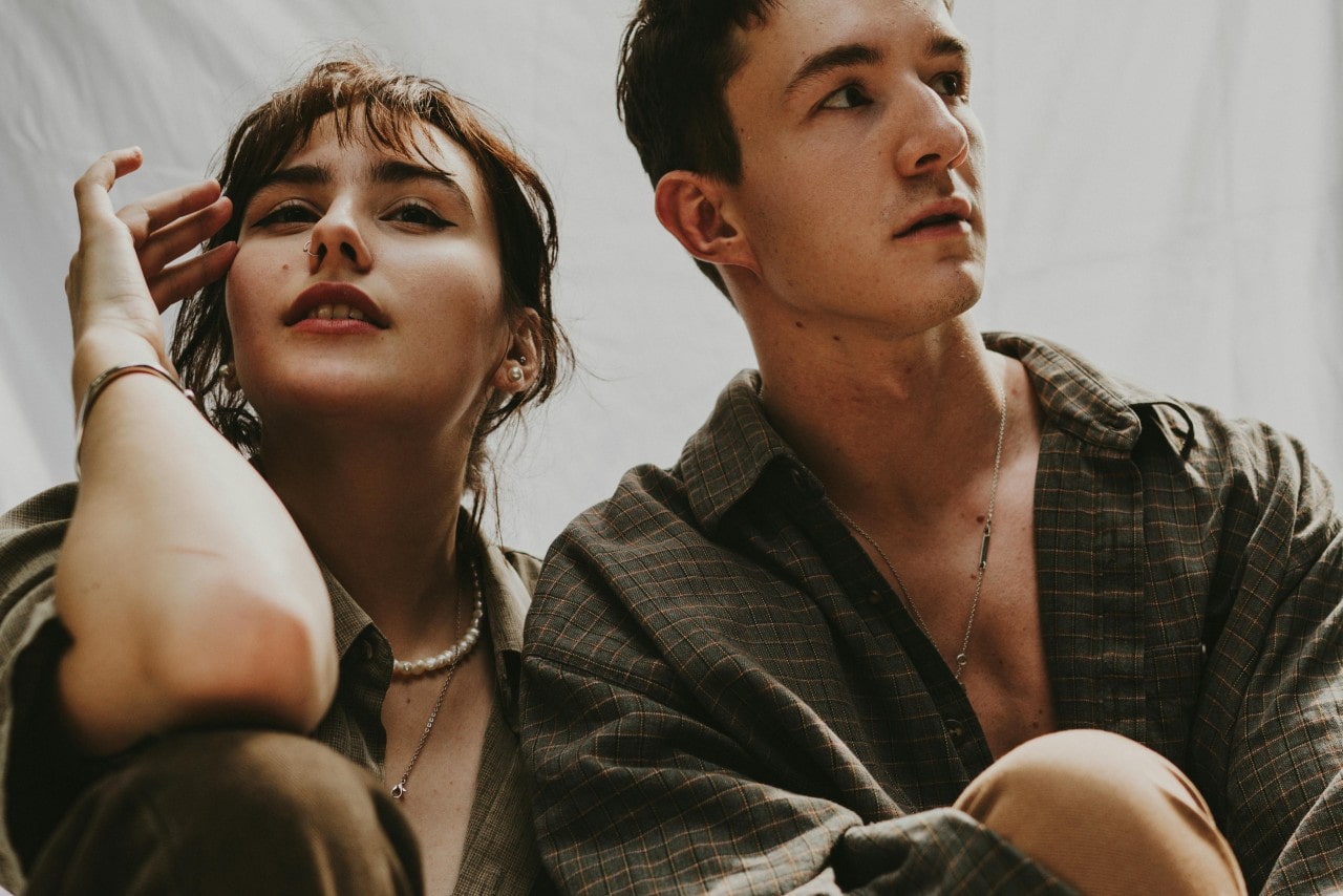 A couple wearing similar green, long sleeve button up shirts. The woman has a pearl necklace with matching pearl stud earrings along with a heart pendant necklace and gold bangle, and the man is wearing a long necklace with bezel set gems along the chain