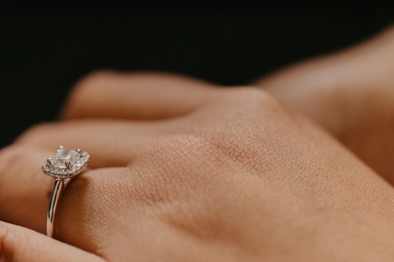 A close-up of a woman’s hand with a halo ring set with a princess cut diamond