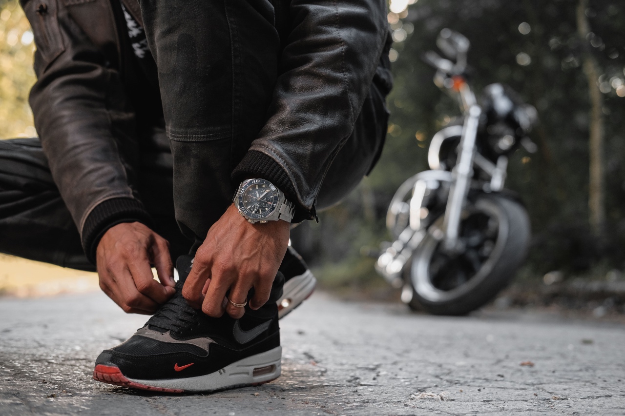 A person tying a black Nike sneaker, showcasing a luxury TAG Heuer chronograph watch with a motorcycle blurred behind.
