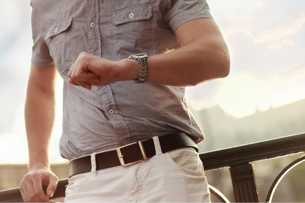 A man in a grey shirt and white trousers checks the time on his stainless steel wrist watch.