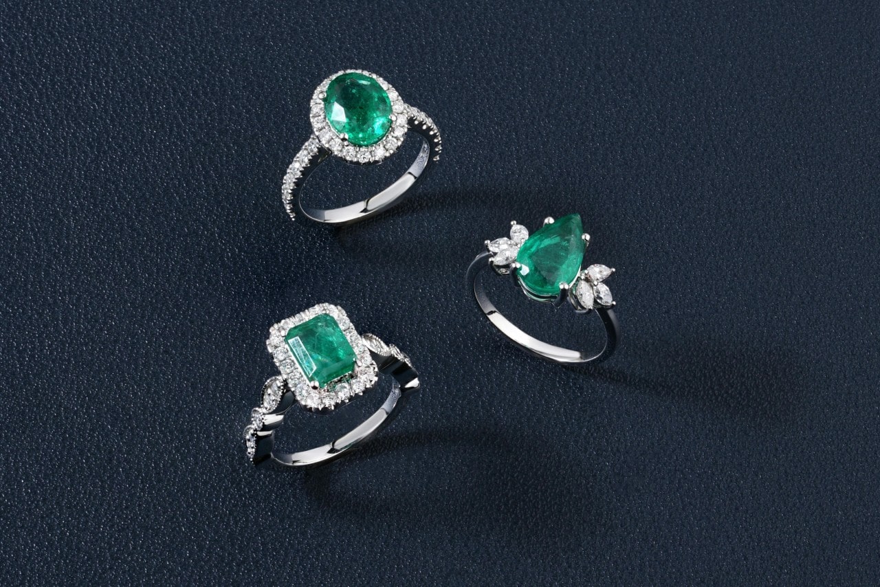 Three white gold rings with vibrant green emeralds and sparkling diamonds on a dark textured surface.