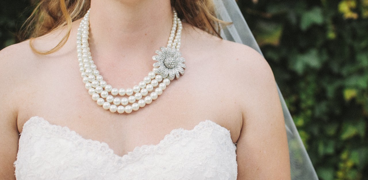 A bride in a strapless lace gown wears a layered pearl necklace with a floral brooch, standing against a leafy green backdrop.