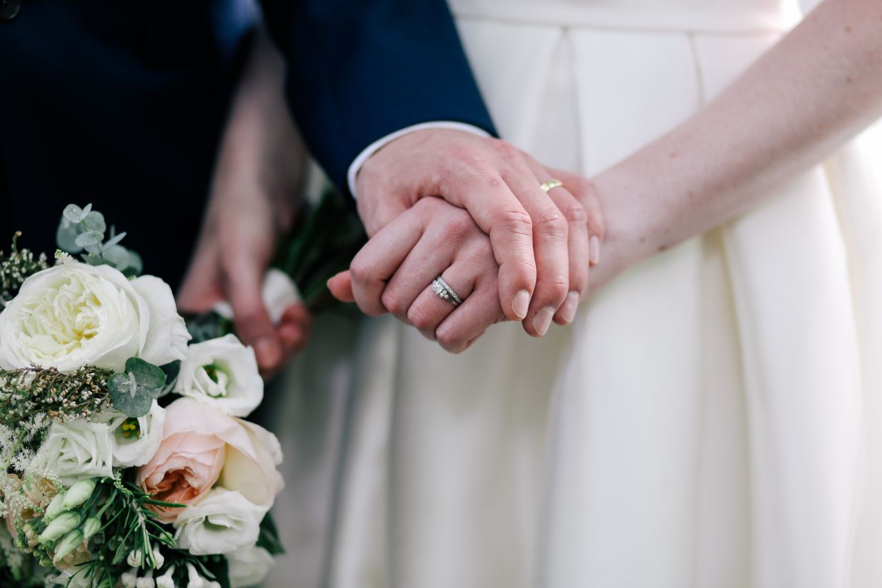 Close-up of a bride and groom holding hands, showcasing wedding rings, while the bride holds a bouquet of white and pink flowers.