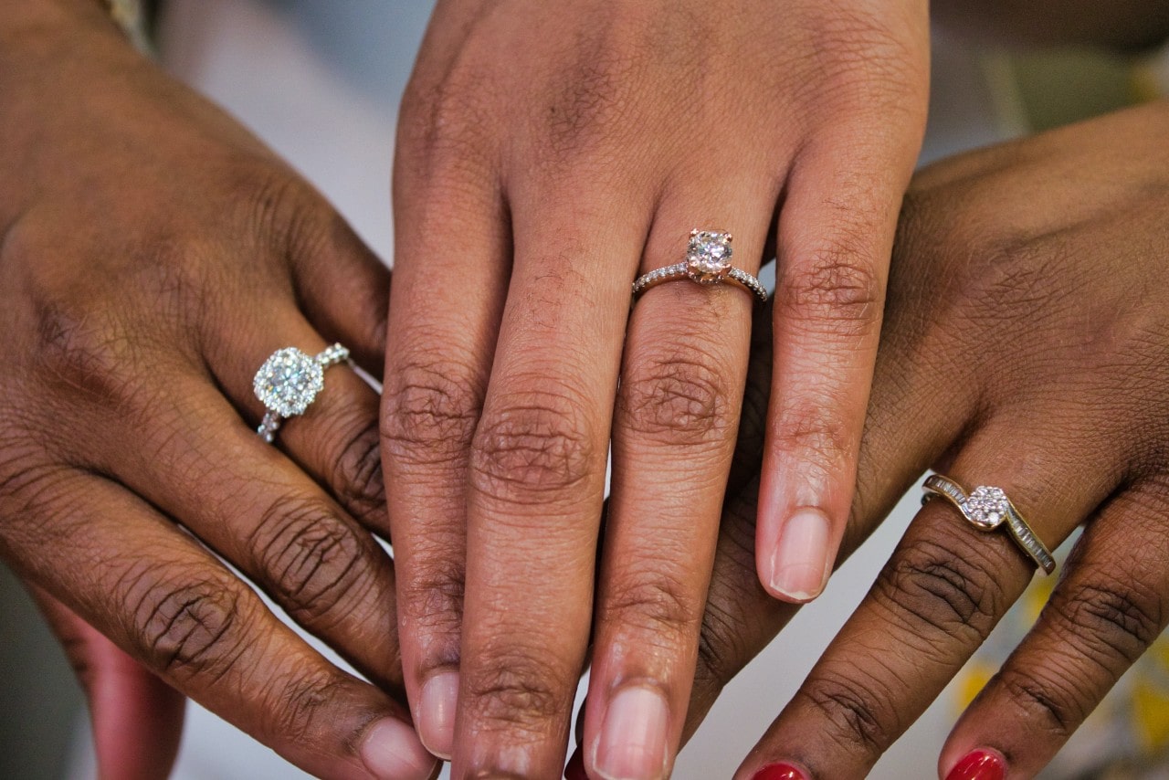 Three women showing off their engagement rings: a halo princess cut with diamonds along the band, a cushion side stone ring, and a round cut with channel set emerald cut stones