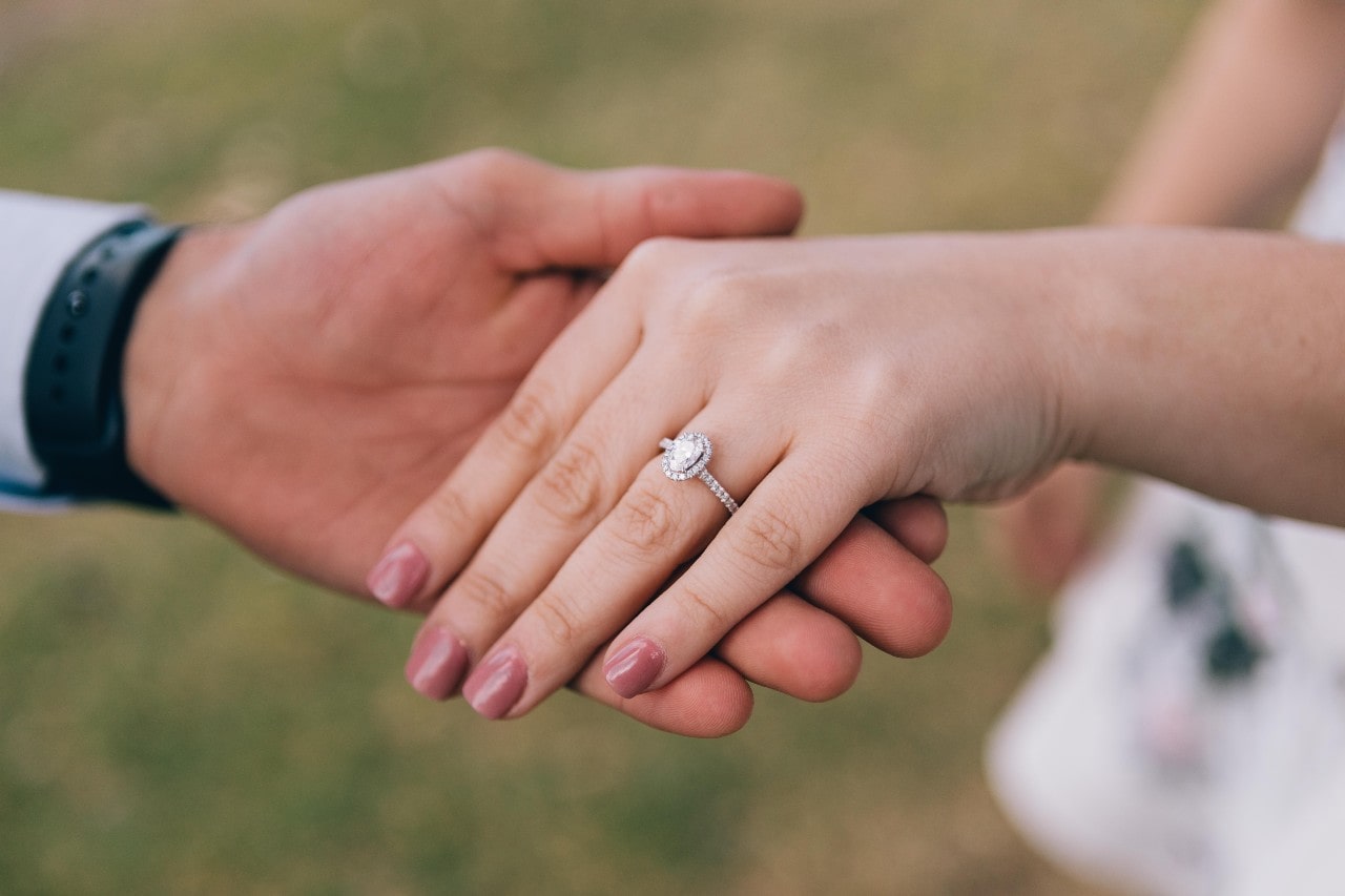 A man holding the hand of a woman with an oval cut halo ring with dainty diamonds all along the band