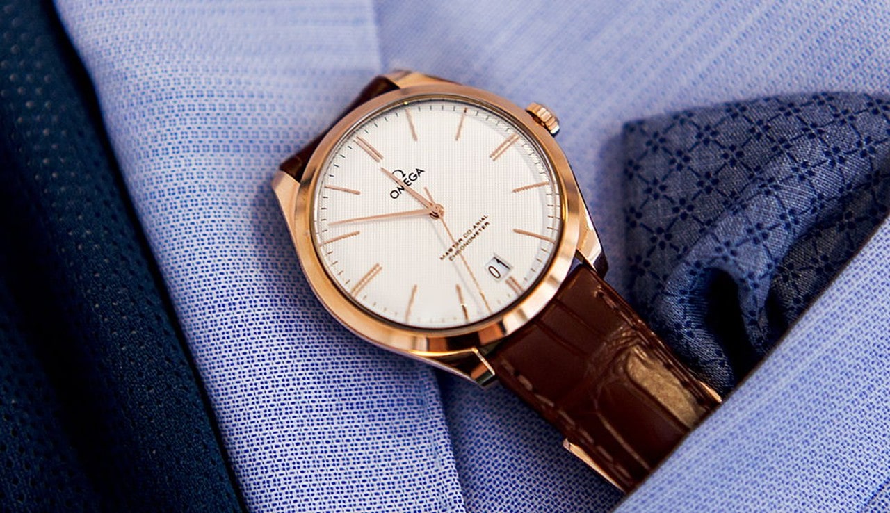 An Omega timepiece with a copper-toned case sits in a man's coat pocket with a handkerchief