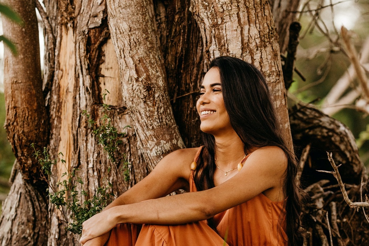 A woman wearing an orange romper and white gold necklace sits under a tree.