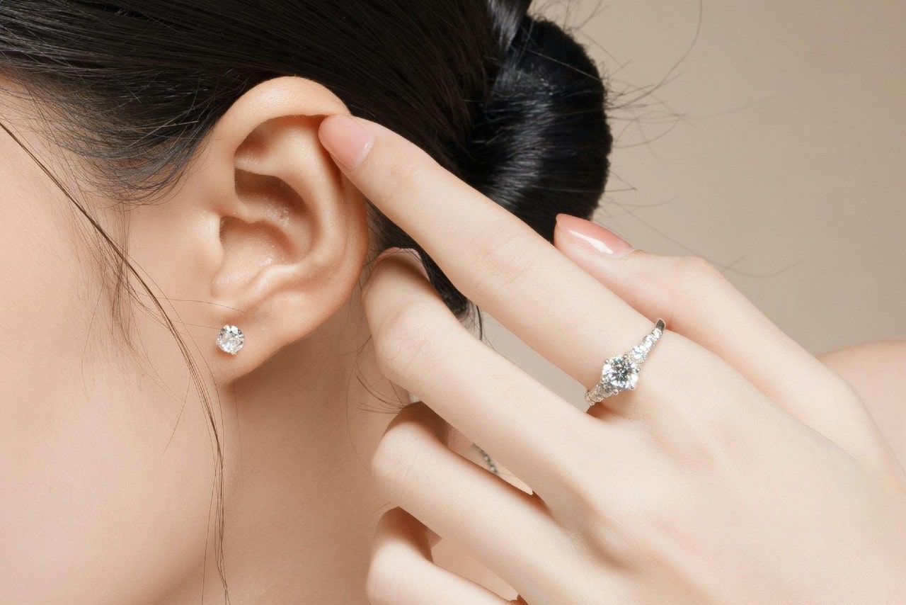 A close-up of a young woman’s hand and ear, her finger adorned with an elegant diamond ring and her ear in simple diamond stud earrings.